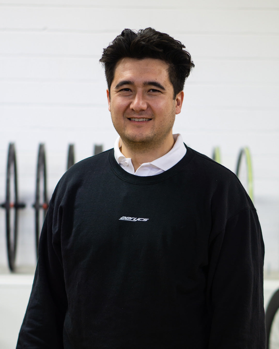 Anthony mit einem aerycs Sweatshirt mit einer weißer Wand und Fahrradreifen im Hintergrund